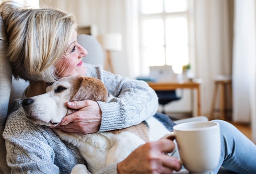 Woman in a cozy sweater hugging a dog while sitting in a bright room, holding a cup of coffee.