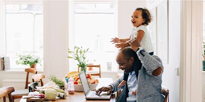 A man with his daughter on his shoulders smiling and using a laptop in a brightly lit room.
