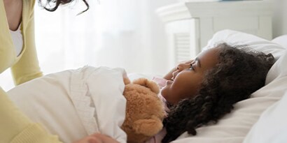 A girl cozy in bed being tucked in with a teddy bear.
