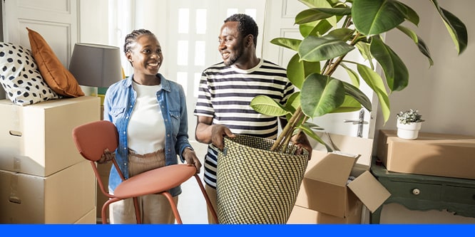 A smiling couple moving into a new home, carrying a chair and a potted plant among stacked moving boxes in a sunlit room.
