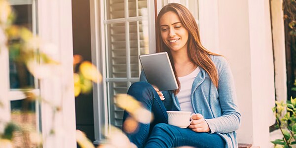 Woman in a blue cardigan sits by a window, reading a tablet and holding a cup, with an expression of contentment.
