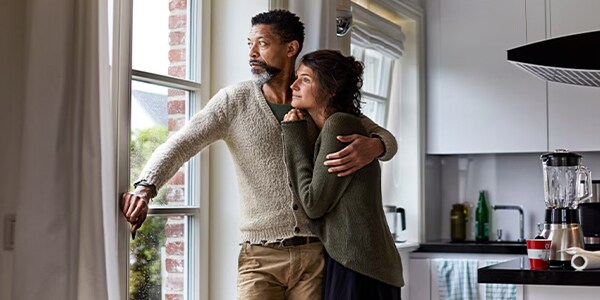 Couple gazes thoughtfully out of a kitchen window, embracing comfortably.
