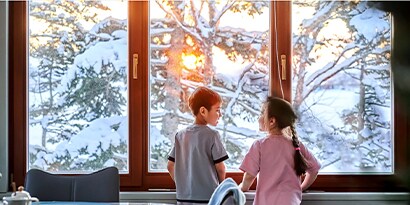 Two children smiling talking while looking out a window at a wintery wonderland.
