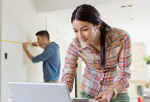 Woman in plaid shirt using laptop while man measures wall in bright room.