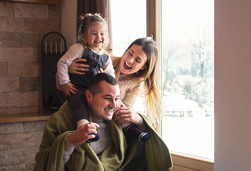 Did you know some of your favorite winter activities may be polluting your indoor air? A father with a child on his shoulders, a mother holding the child smiling. They sit in front of a wintery window.
