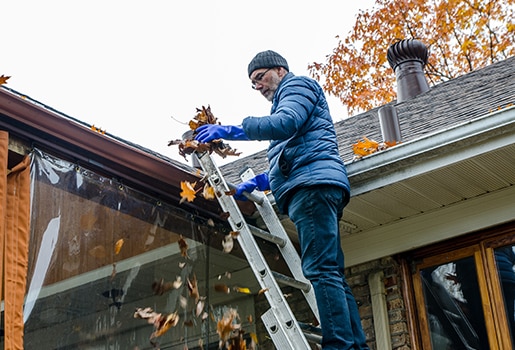 A Man in a puffy navy jacket on a ladder removing leaves from the gutters of a house. Tackle these 10 seasonal chores when daylight savings ends.
