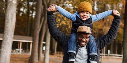 A man with his son on his shoulders both smiling, a fall landscape behind them.
