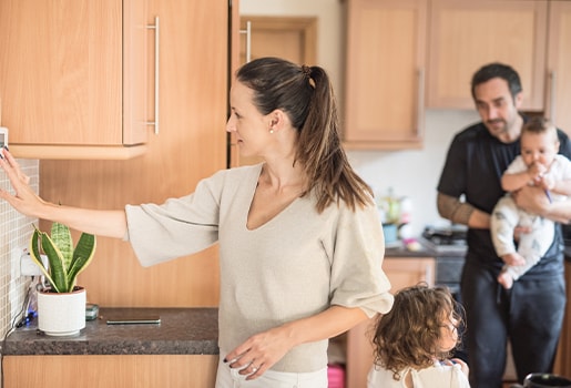 Woman adjusts a thermostat in a kitchen, with a man holding a baby in the background.