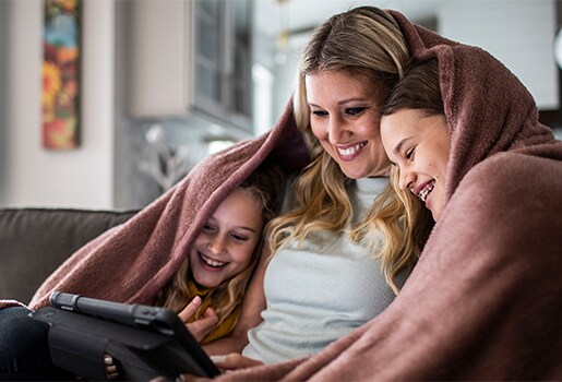 Cozy season is coming! Here’s how to prep your home. Three people on a couch, cozy wrapped in a blanket looking at a tablet.