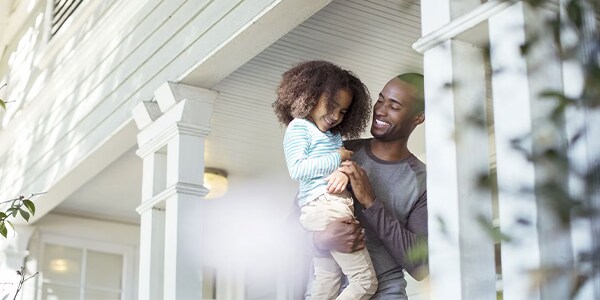 A man smiling holding his child on their front porch.
