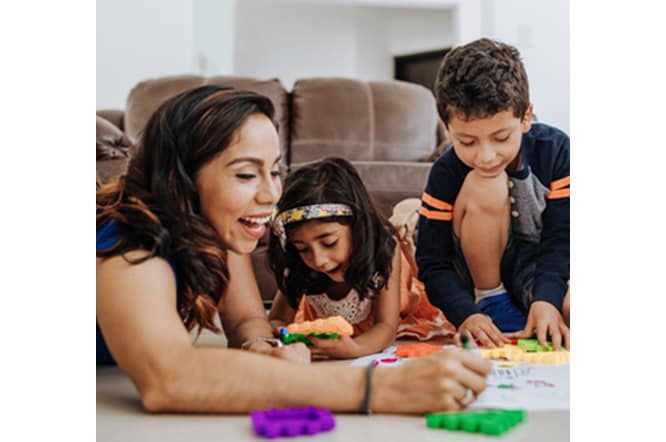 A family sitting on the floor in a living room smiling, doing crafts together.
