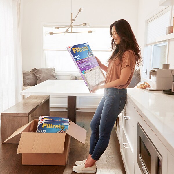 Woman in a kitchen holding a Filtrete air filter, with an open box of additional filters on the floor.
