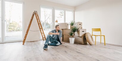 A man sitting in an spacious room with moving boxes, a chair and a ladder.

