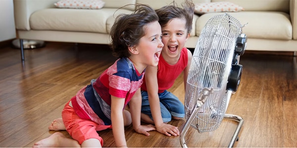 Two children happily play with a floor fan in a living room with wooden flooring and a beige sofa.
