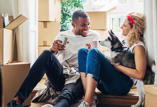 A couple sits among moving boxes, smiling and holding mugs, with a small dog on their lap.