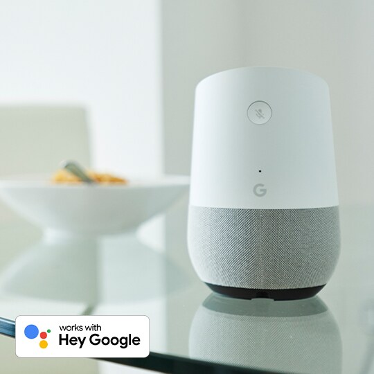 Head on shot of Google Assistant on a glass table. Ceramic bowl of fruit sits out of focus in the background. Works with Hey Google in lower left corner