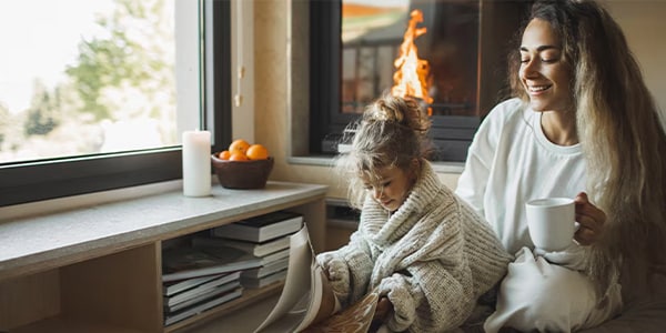 A woman holding a mug and child sitting on the ground reading books in front of a fireplace.
