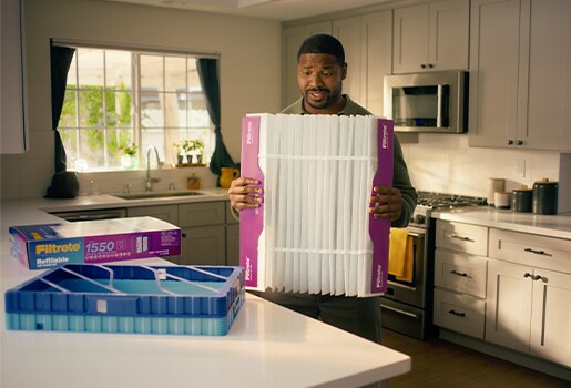 A man in a kitchen holds a large Filtrete™ Refillable Air Filter. Nearby is a Filtrete™ Refillable Air Filter Kit and a blue filter tray.