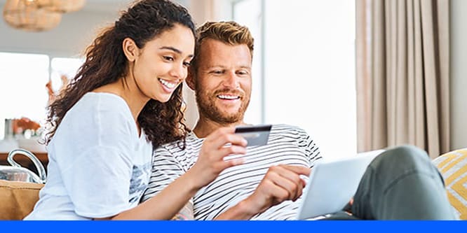 A couple sitting on a couch smiling while looking at a credit card and tablet.
