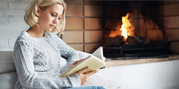 A person in a gray sweater reads a book by a lit fireplace, creating a cozy atmosphere.
