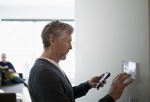 Man with phone in left hand, while adjusting a smart thermostat in a brightly lit room.