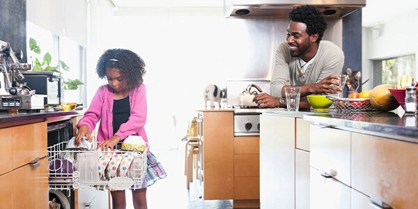 Girl unloads a dishwasher while a man watches and smiles in a modern kitchen.
