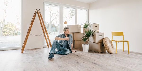 Man sitting on floor with coffee, surrounded by moving boxes, ladder, and yellow chair in a bright room.
