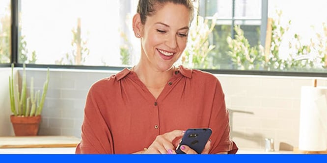 A smiling woman wearing a red button up shirt looking at her black smart phone.
