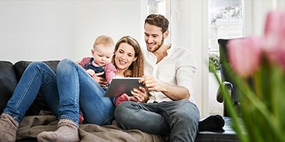 A mother and father with a baby on a couch smiling at a tablet.
