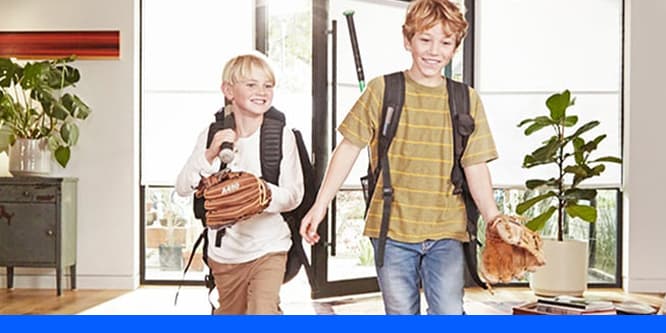 Two smiling kids coming inside a house with holding backpacks and baseball gloves.
