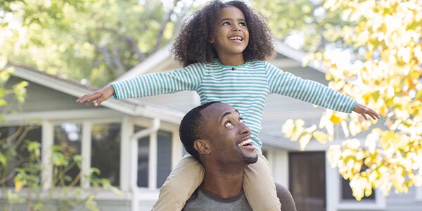 Child with curly hair sits on an adult's shoulders, both smiling, outside a house with autumn trees in the background.
