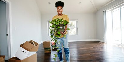 A woman holding a house plant in an empty room with moving boxes on her left.
