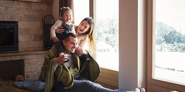 A family of three enjoys a cozy moment by a window on a snowy day, with a girl on her dad's shoulders.
