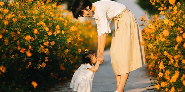 A woman bends to embrace a child in a pathway surrounded by blooming orange flowers under sunlight.
