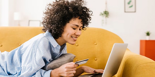 Person smiling, holding a credit card while using a laptop on a yellow couch at home.
