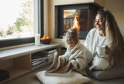 A mother and daughter reading a book together in front of a fireplace. Discover how to enhance your home’s coziness this winter with candle and fireplace best practices and alternatives that prioritize air quality.