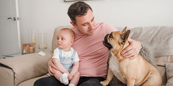 A father sits on a couch with his dog and baby on his lap.
