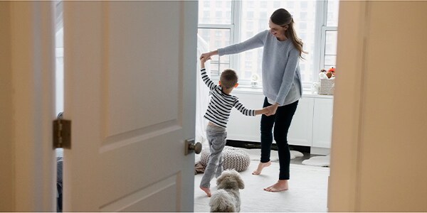 A woman and child dance in a living room with a dog, viewed through an open door.
