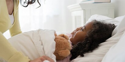 A mother tucking in a smiling child into bed with a teddy bear.
