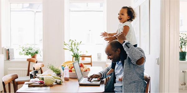 Man working on a laptop at a kitchen table with a child on his shoulders, surrounded by plants and groceries.

