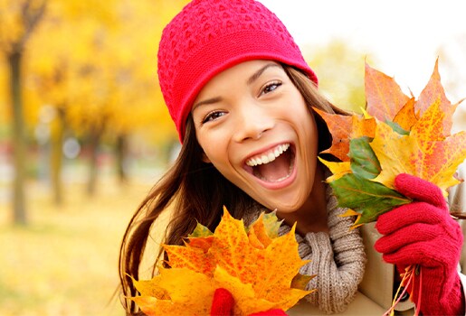Woman in a red hat and gloves smiling, holding autumn leaves with vibrant yellow trees in the background.