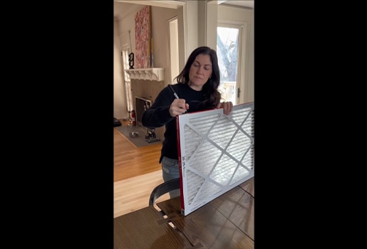 Person in a living room writes on an air filter with a marker, near a fireplace and a large window.