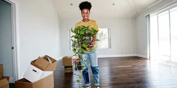 Person smiling and holding a potted plant, standing in a bright, empty room with unpacked moving boxes.
