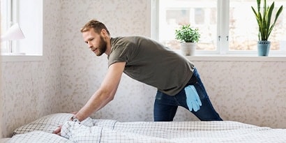 A man making the bed in a well lit bedroom.
