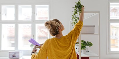 A woman in a yellow shirt placing a plant on a high shelf.

