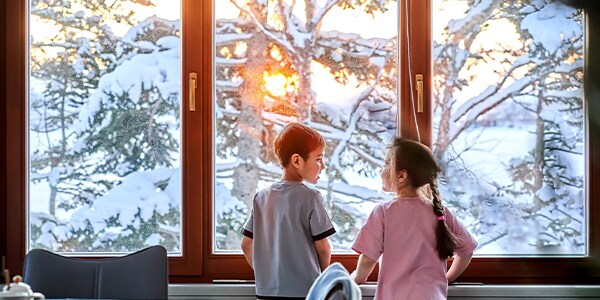 Two children looking out a window at a snowy landscape with sunset.
