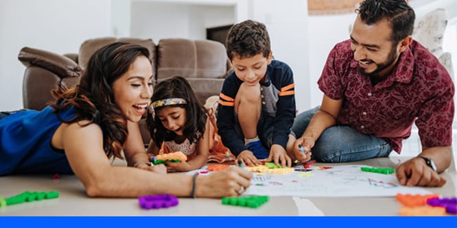 A Family of four sitting on the floor in a living room smiling, doing crafts together.
