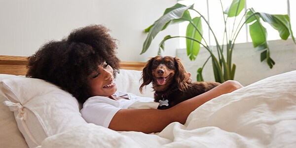 A person smiles while lying in bed, cuddling a happy dog, with green plants in the background.
