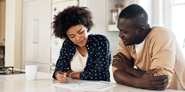 Two people sit at a kitchen table reviewing documents together with a mug nearby.
